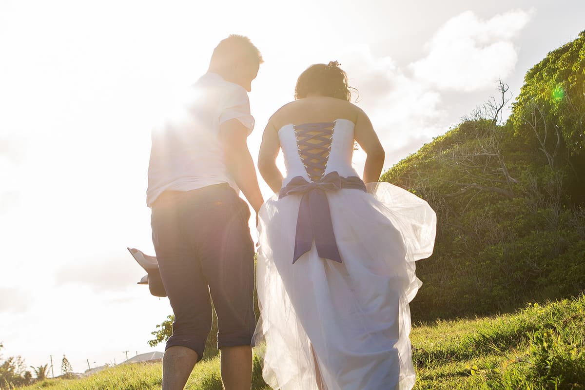 Bride and groom walking barefoot up a grassy hill into the sunlight in Barbados