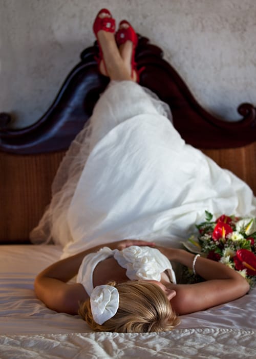 Bride resting on a bed in her wedding dress with red shoes and bouquet