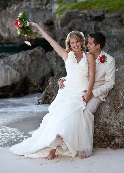 Smiling bride raising her bouquet while seated with the groom by the sea