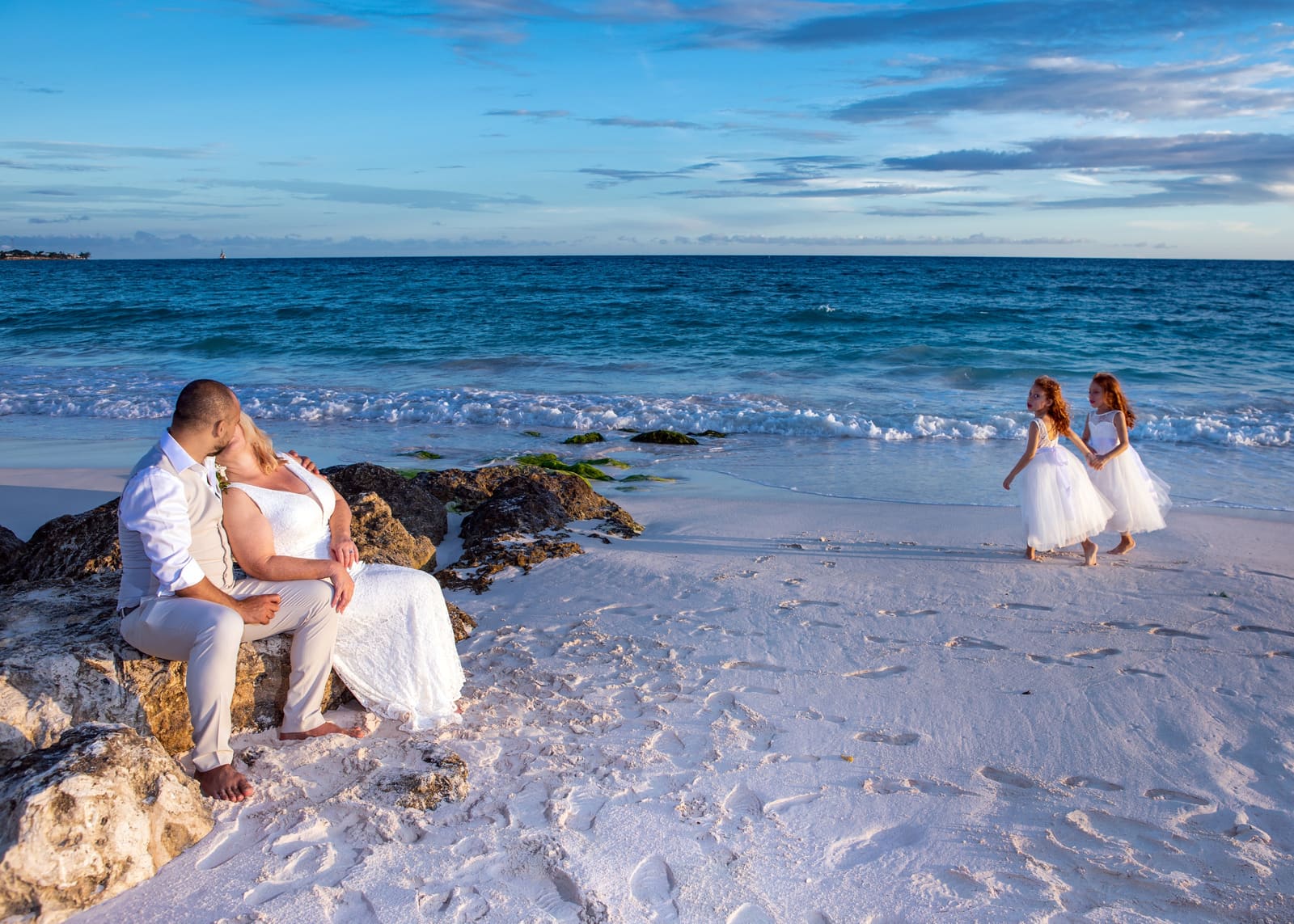 Couple seated on rocks by the sea as two flower girls play on the sand, Barbados